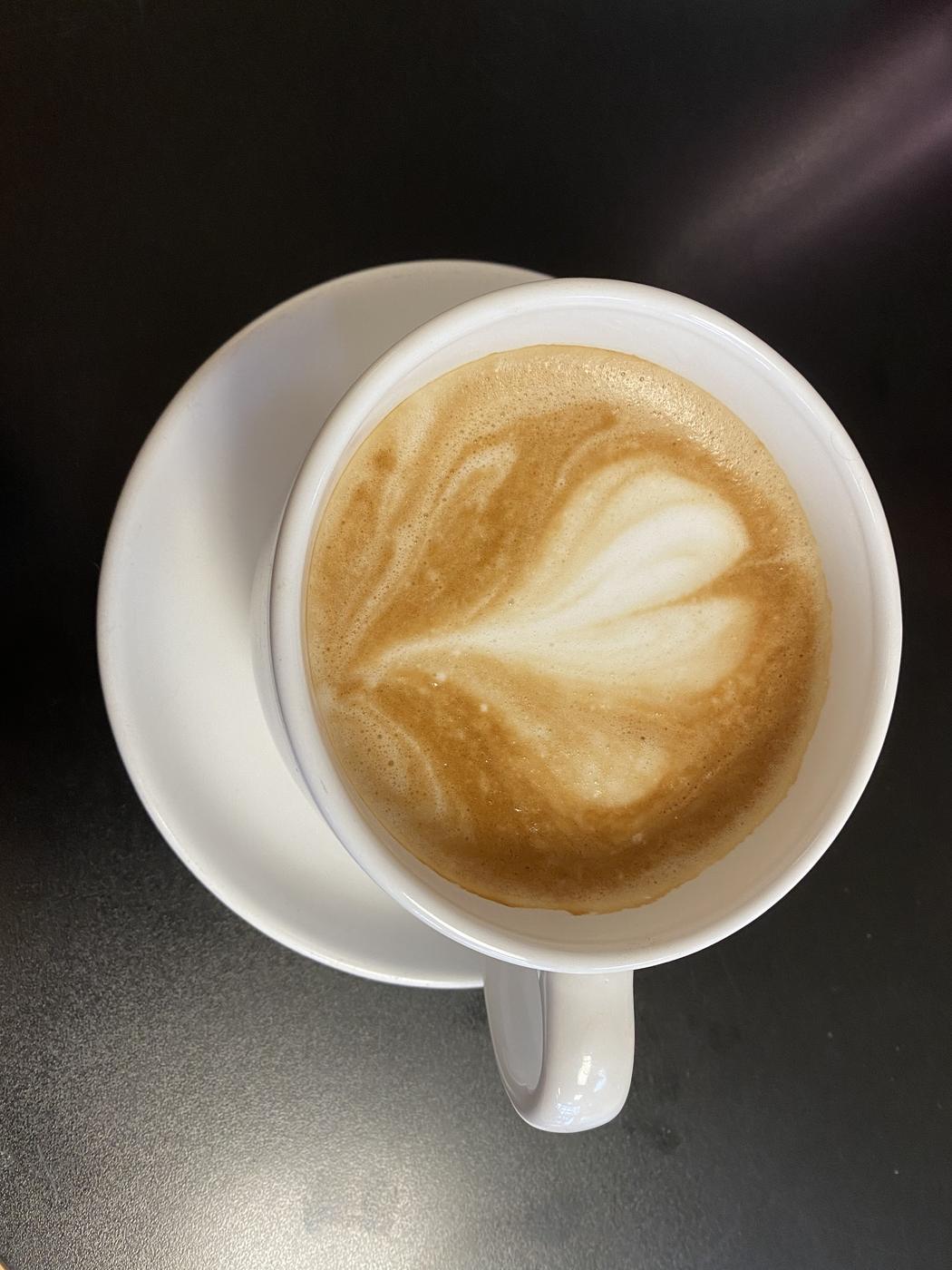 A latte with rosetta art in a white cup and saucer on a dark table, seen from above.